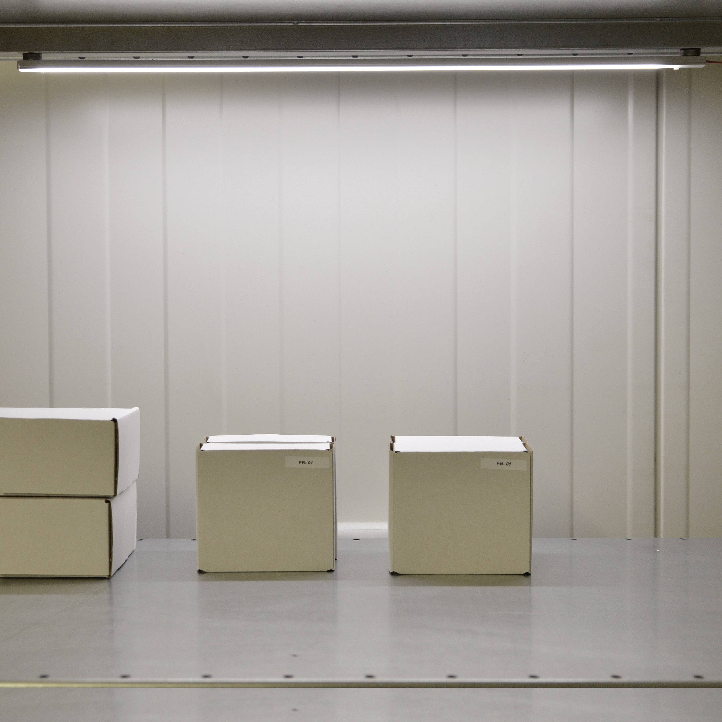 Four white boxes on a metal shelving unit in a storage room. Three stand individually side by side, one stacked. Overhead lighting illuminates the space.
