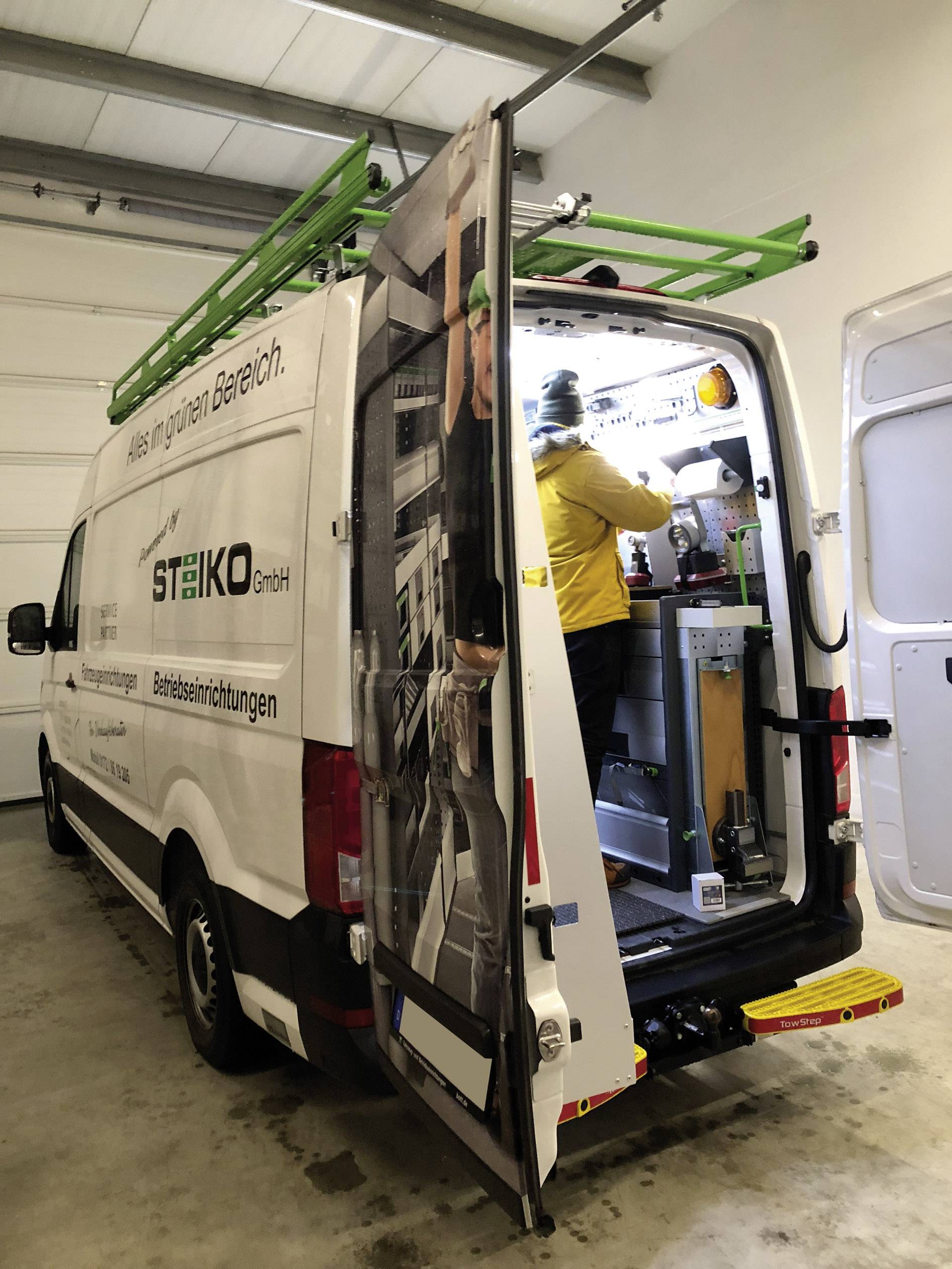 A tradesman is working in an open van with tools and equipment inside. The van has a ladder on its roof.