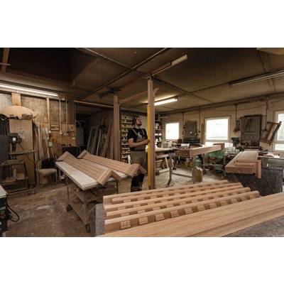 A craftsman is working in a woodwork workshop, surrounded by wooden boards and tools. Daylight streams in through the windows.