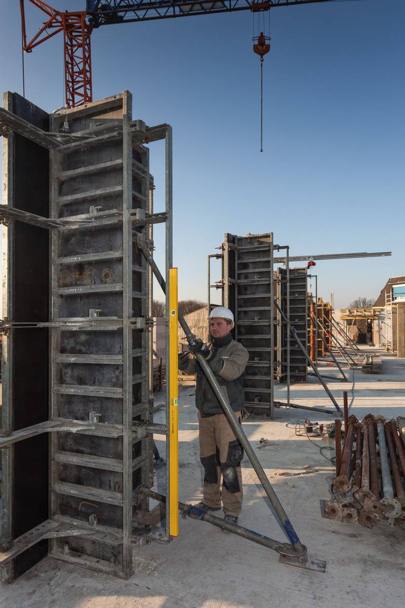 A person in work attire is working on a construction site. They are measuring a standing metal formwork with a yellow spirit level. Cranes and additional formwork can be seen in the background.