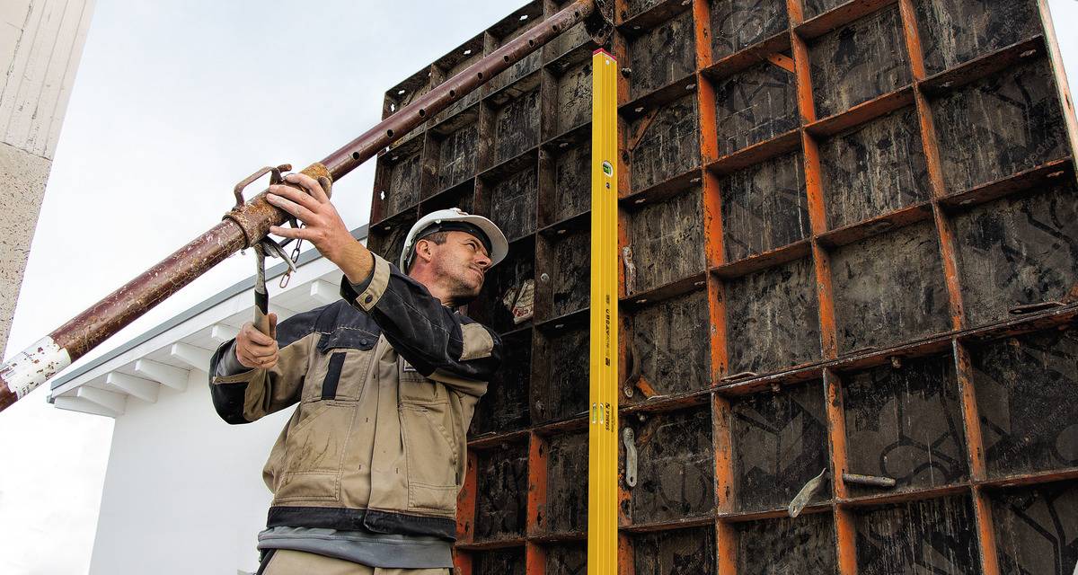 A construction worker is aligning a construction support on a large formwork wall while wearing a hard hat. A spirit level is positioned nearby.