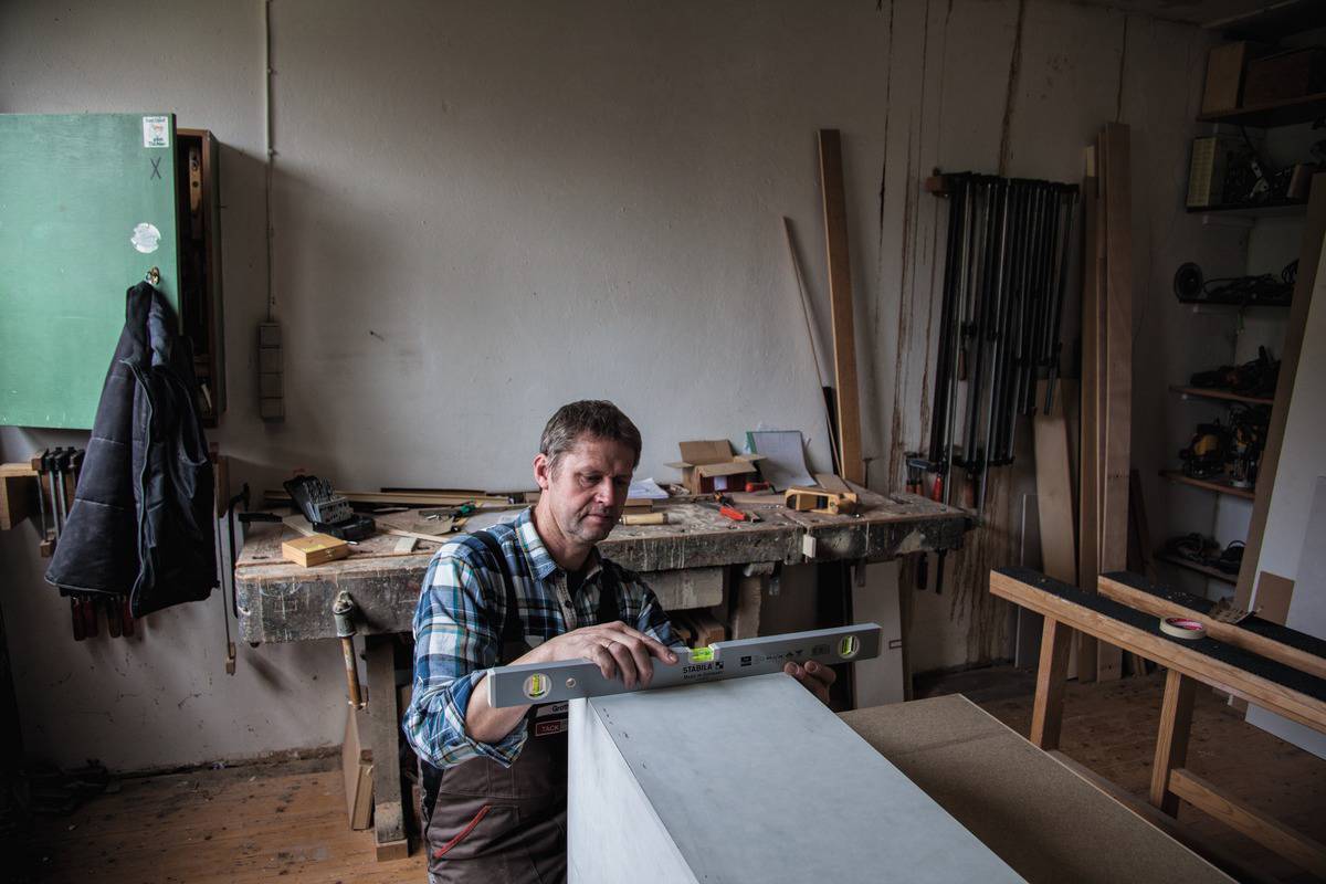A man in a workshop is using a spirit level to check the surface of a light-coloured board. Tools can be seen in the background.