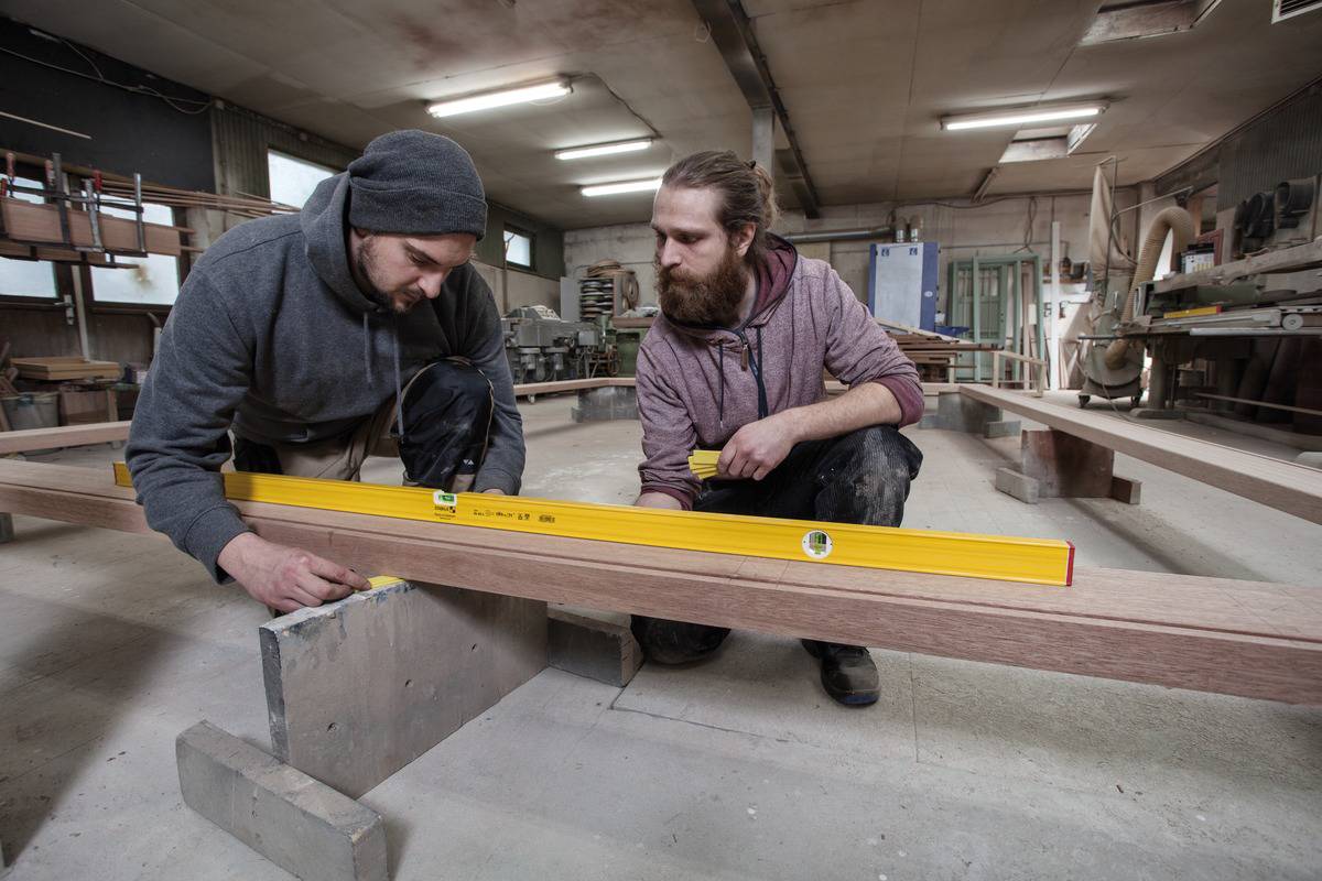 Two men in a workshop are using a yellow spirit level to measure and align a long wooden board on a workbench.