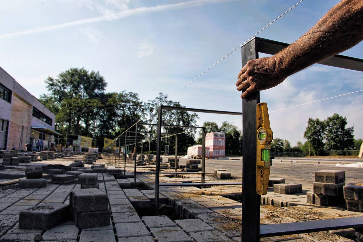 A construction site with a worker aligning a scaffold. Buildings and trees can be seen in the background.