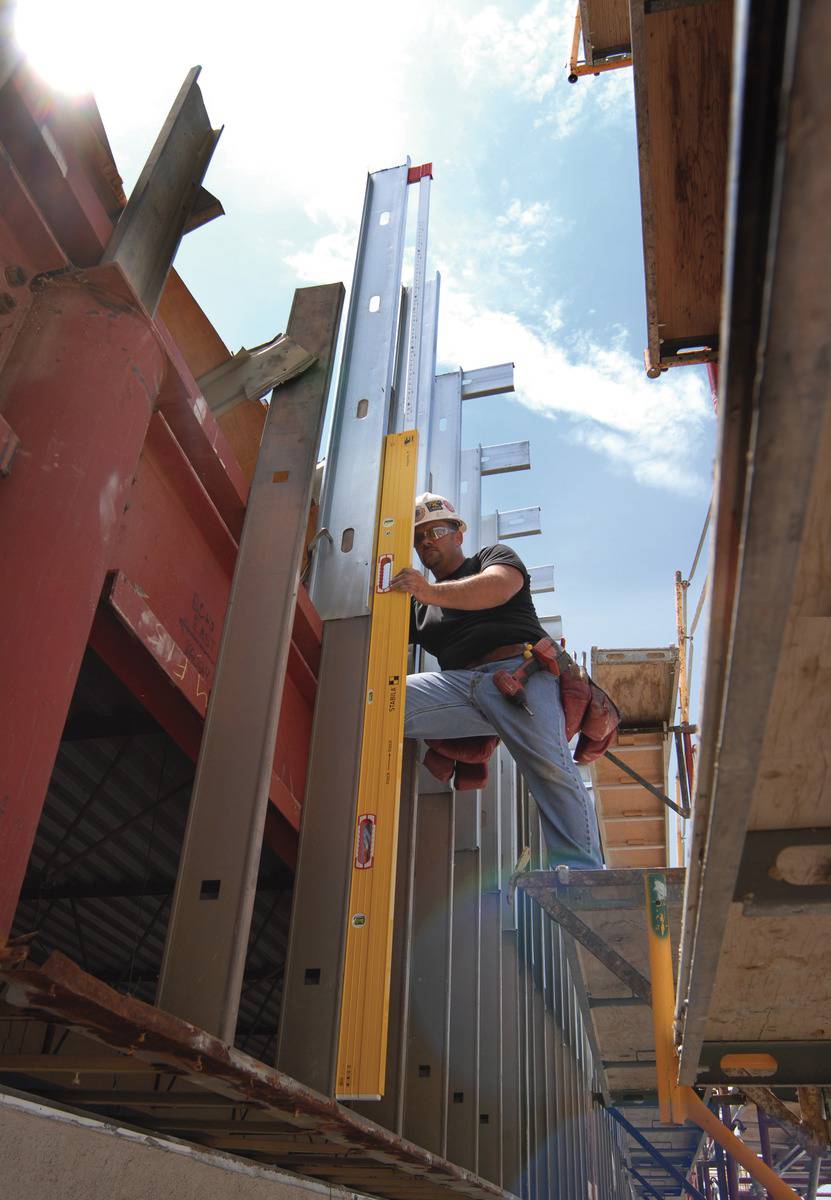 A worker on a construction site, high up on scaffolding, is using a spirit level to align metal beams on a building structure.
