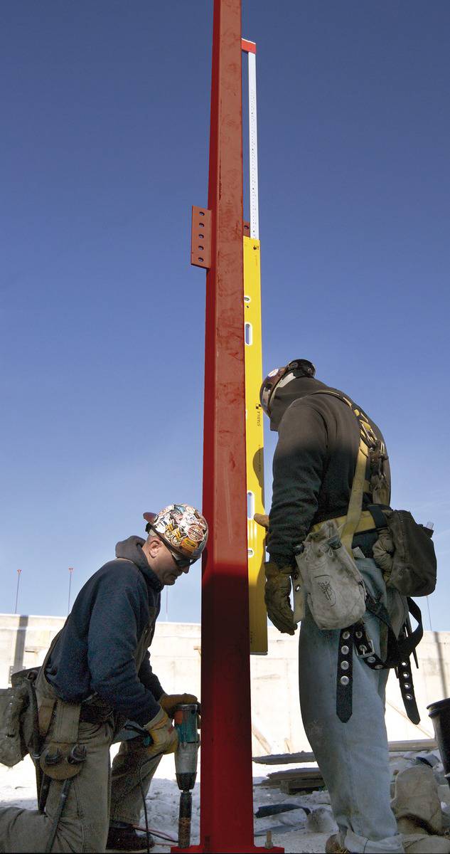 Two construction workers are installing a red steel support. One is holding a spirit level, the other is using a drill. Background: clear sky.