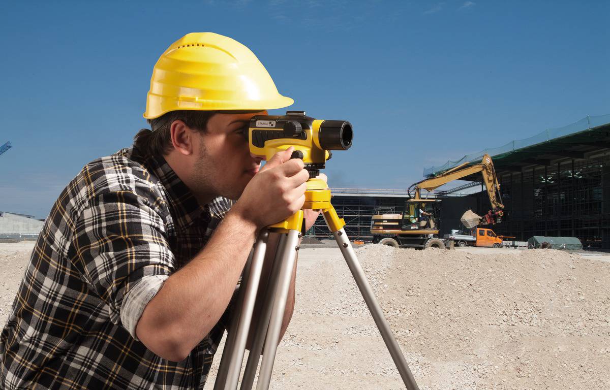 A construction worker wearing a yellow hard hat is using a surveying instrument on a building site. A digger can be seen in the background.