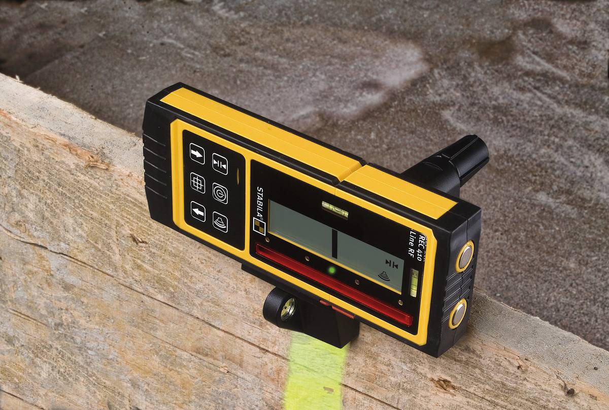 A yellow laser measuring device with a digital display is attached to a wooden board and is measuring the inclination. The background shows a rough surface.