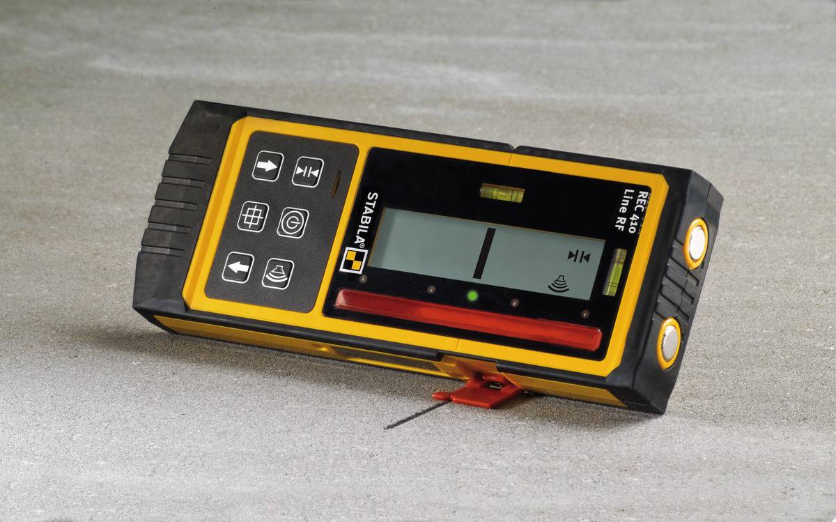 An electronic laser receiver is resting on a grey surface. The device displays symbols and buttons for operation.