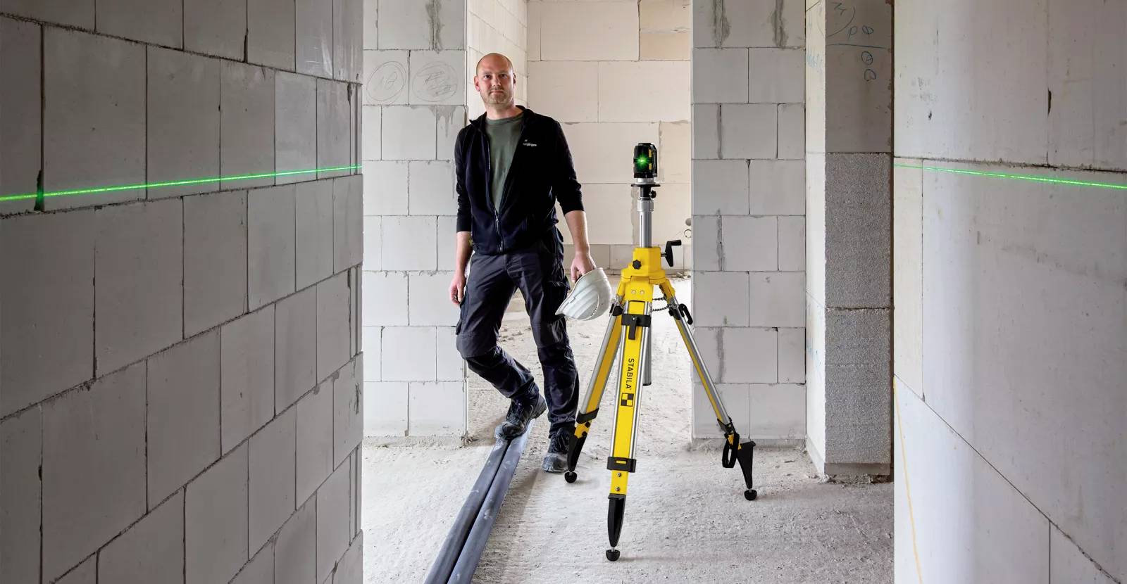 A person holding a bucket stands beside a yellow laser level tripod in a partially constructed room with cinderblock walls marked by green laser lines.