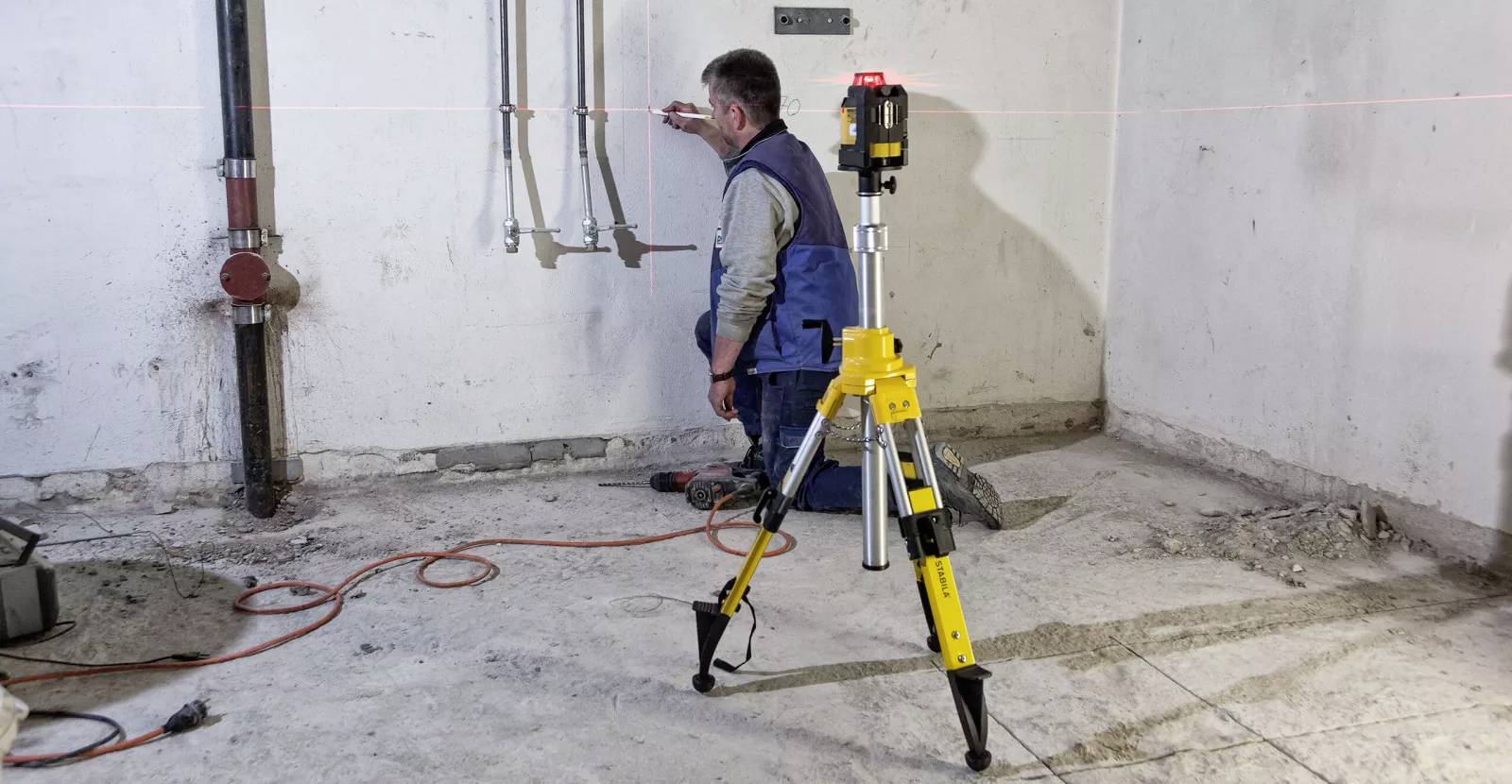 A man kneels on a construction site floor, using a laser level placed on a tripod for accurate pipe installation alignment on a wall.