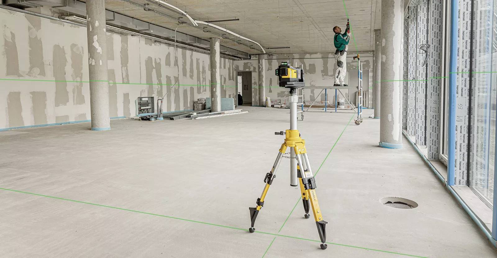 A construction worker on scaffolding installs fixtures in a large, unfinished room. A laser level projects green lines on the floor and walls.