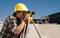 A civil engineer wearing a yellow hard hat is using a surveying instrument on a construction site, with an excavator in the background.