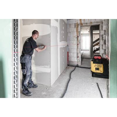 A construction worker is measuring distance with a red laser on a plasterboard wall in an unfinished room. A yellow device is standing nearby.