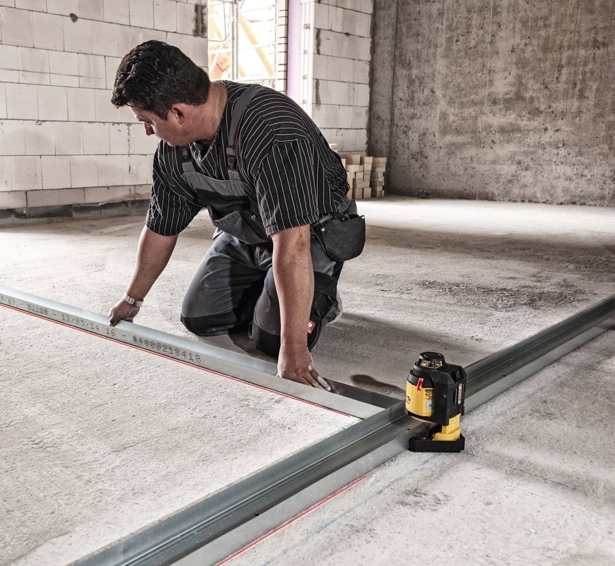 A man is aligning metal strips on a concrete floor, assisted by a yellow laser tool for precise positioning.