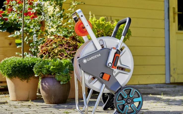 A portable garden hose reel with a handle and wheels is positioned on a brick patio near vibrant potted plants against a yellow wall.