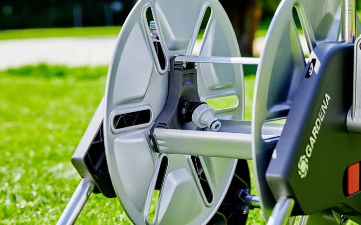 A close-up of an empty, silver hose reel on a lush green lawn, showcasing the brand 'Gardena' on the side.