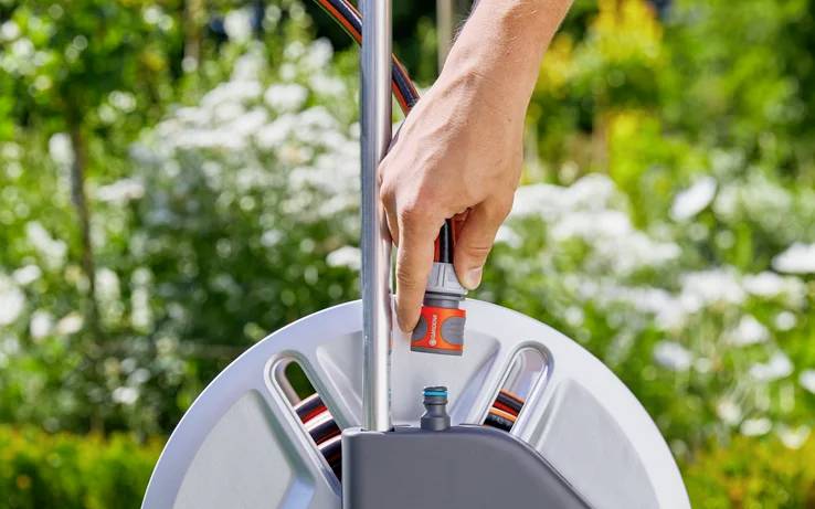 A hand attaches a garden hose to a reel in a lush garden, surrounded by blooming white flowers and greenery, suggesting garden maintenance.
