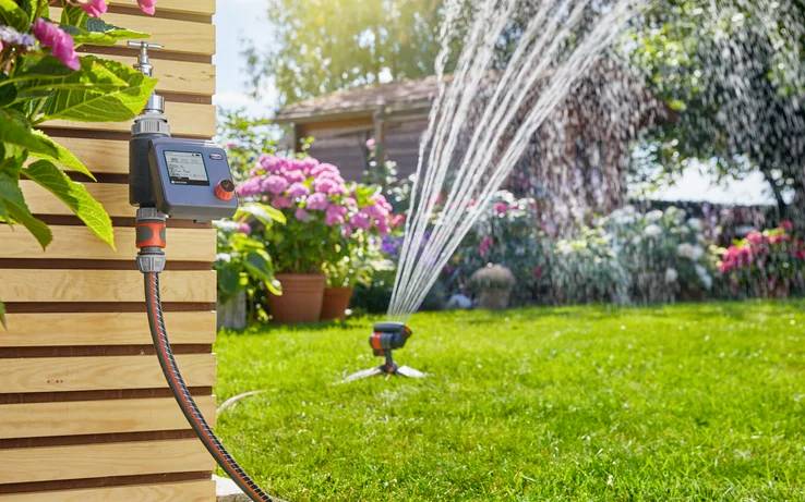 A garden with a sprinkler watering the grass; a digital timer is attached to a wooden wall, controlling the irrigation system.