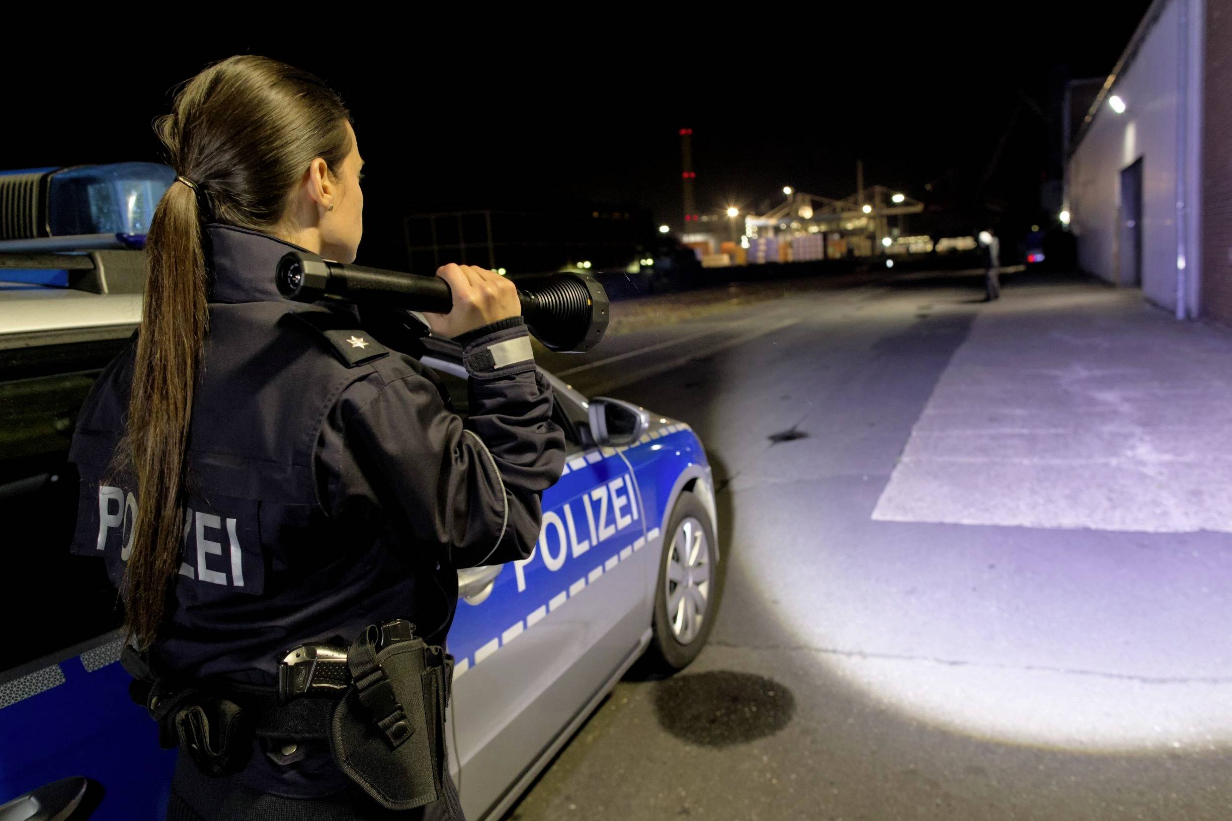 A police officer stands beside a police vehicle at night, shining a torch into a dark area.