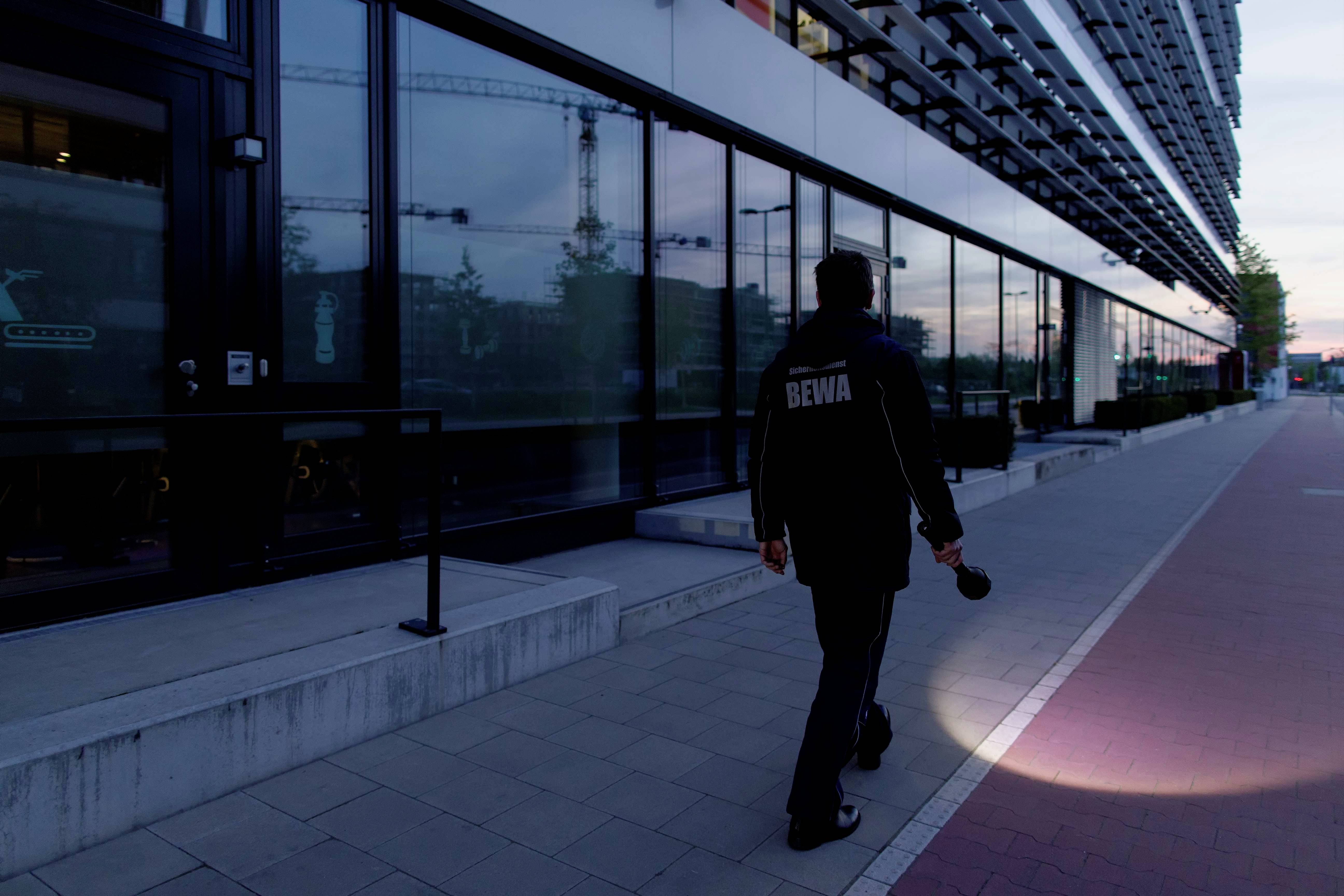 A security guard walks along a pavement with a torch, beside a modern building at dusk.