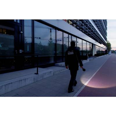 A security guard walks along a pavement with a torch, beside a modern building at dusk.