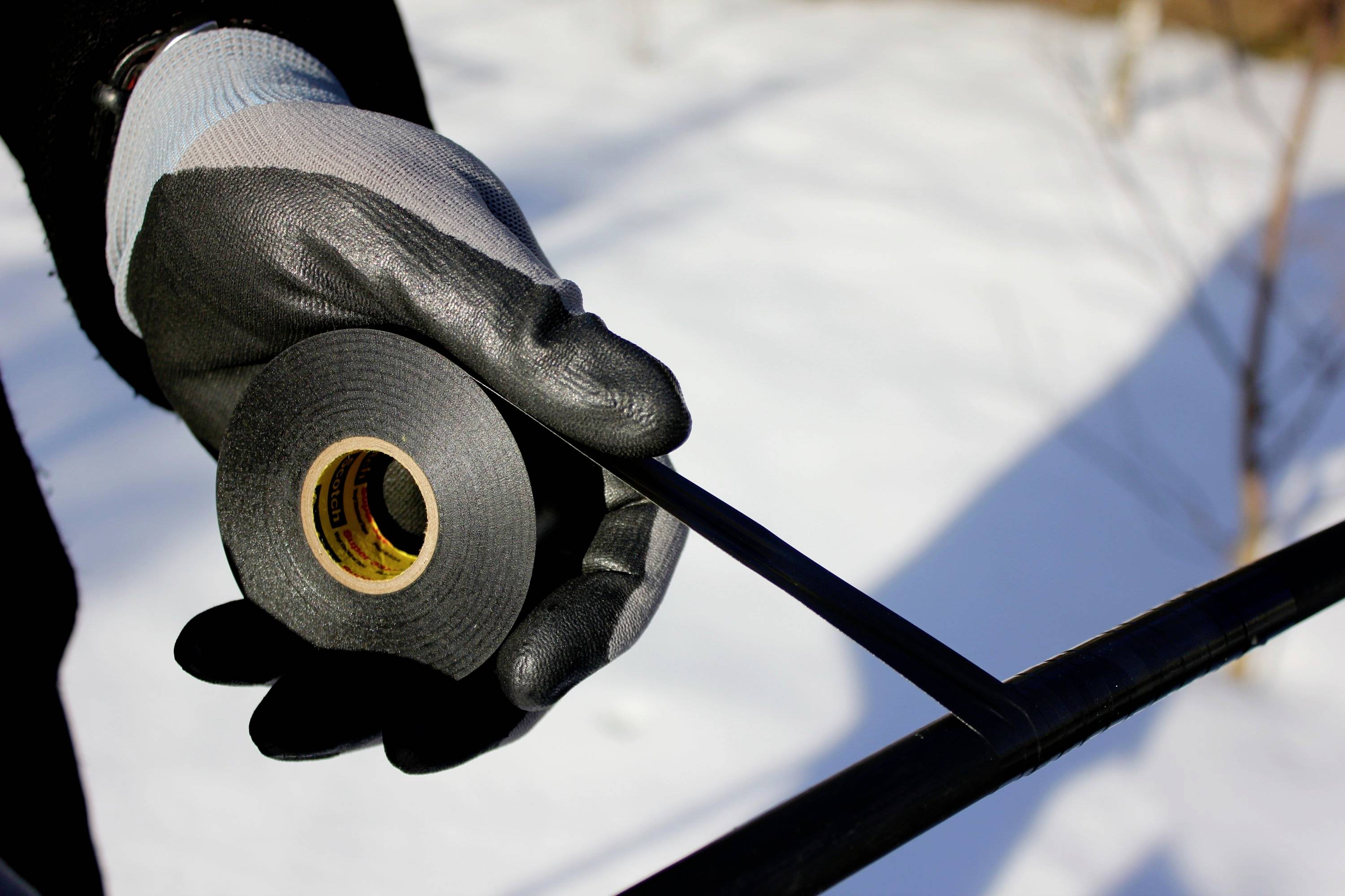 A gloved hand holds insulating tape and wraps a cable outdoors. The background shows snow.