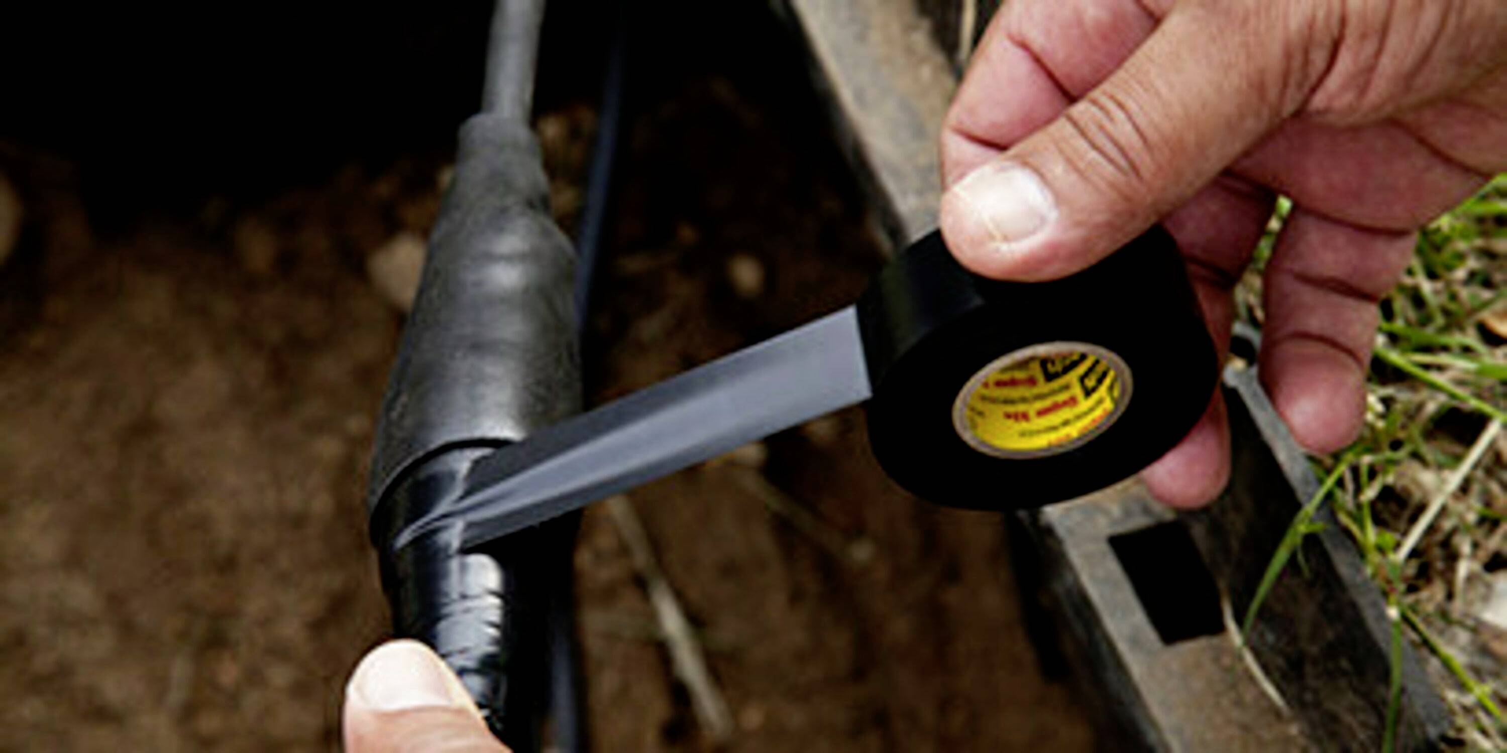 'Close-up of hands wrapping black electrical tape around a cable lying outdoors in the ground to secure it.'