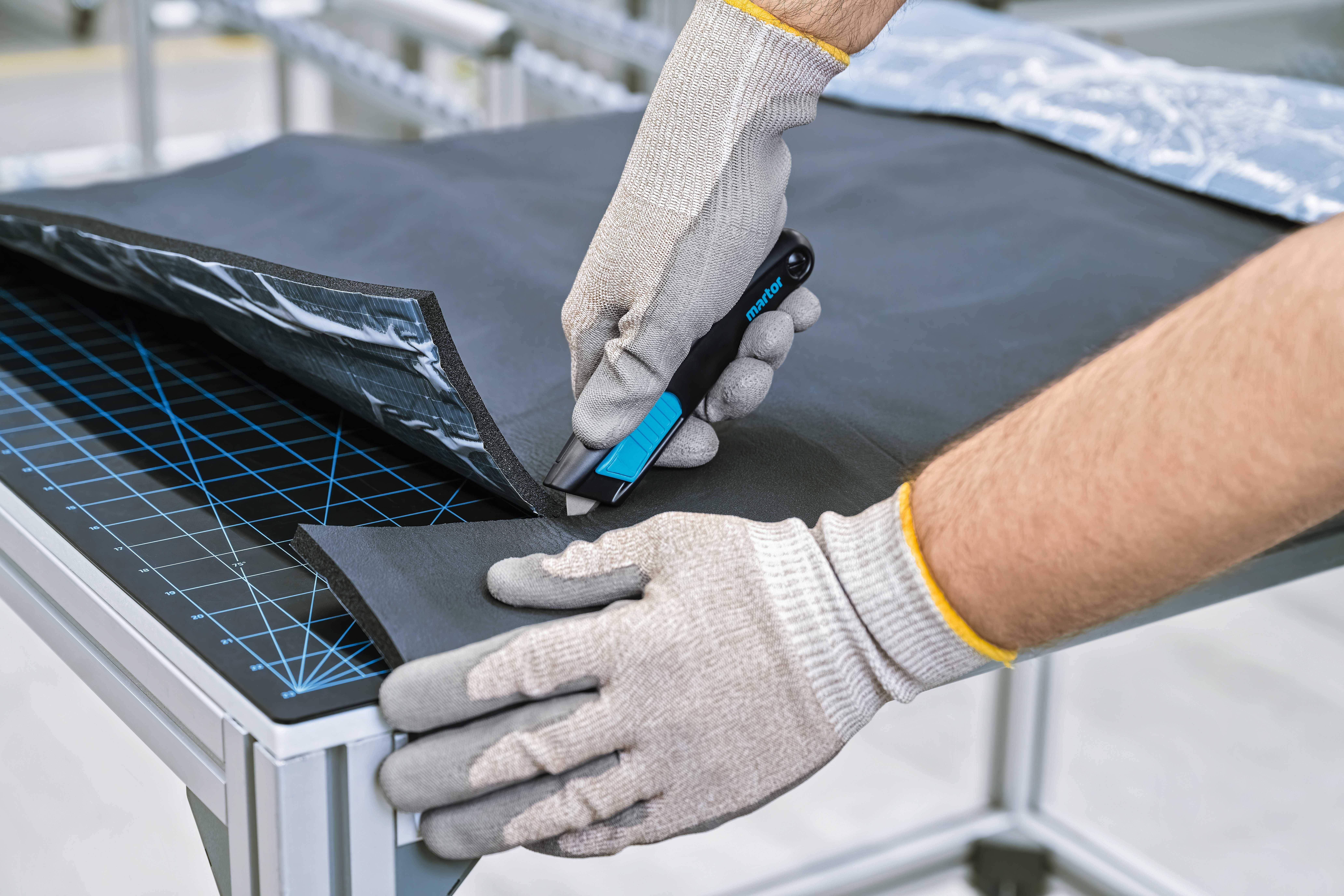 A person is cutting a black material with a blue craft knife on a table, wearing protective gloves.