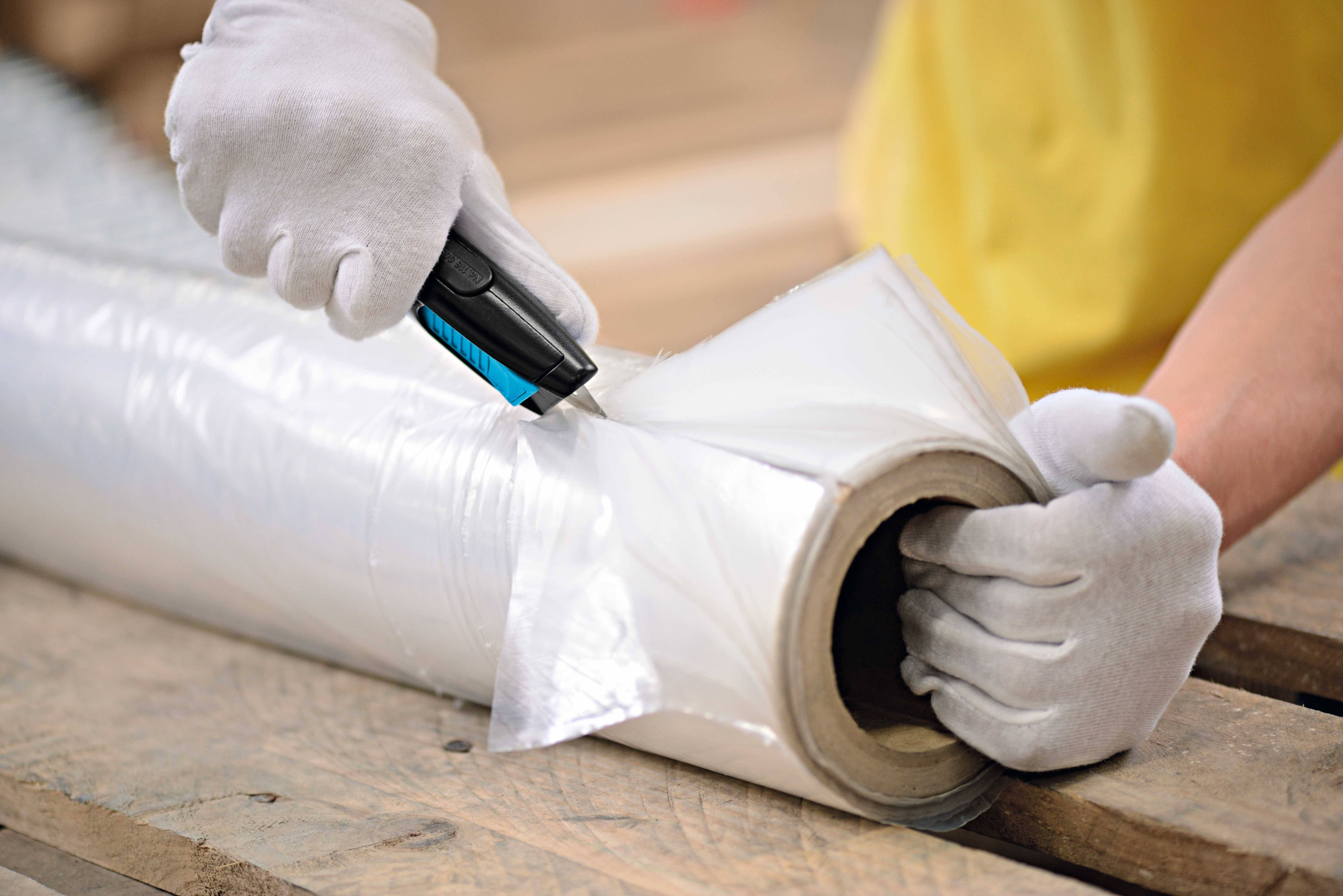 A person wearing white gloves is cutting plastic film from a roll on a wooden pallet using a knife.