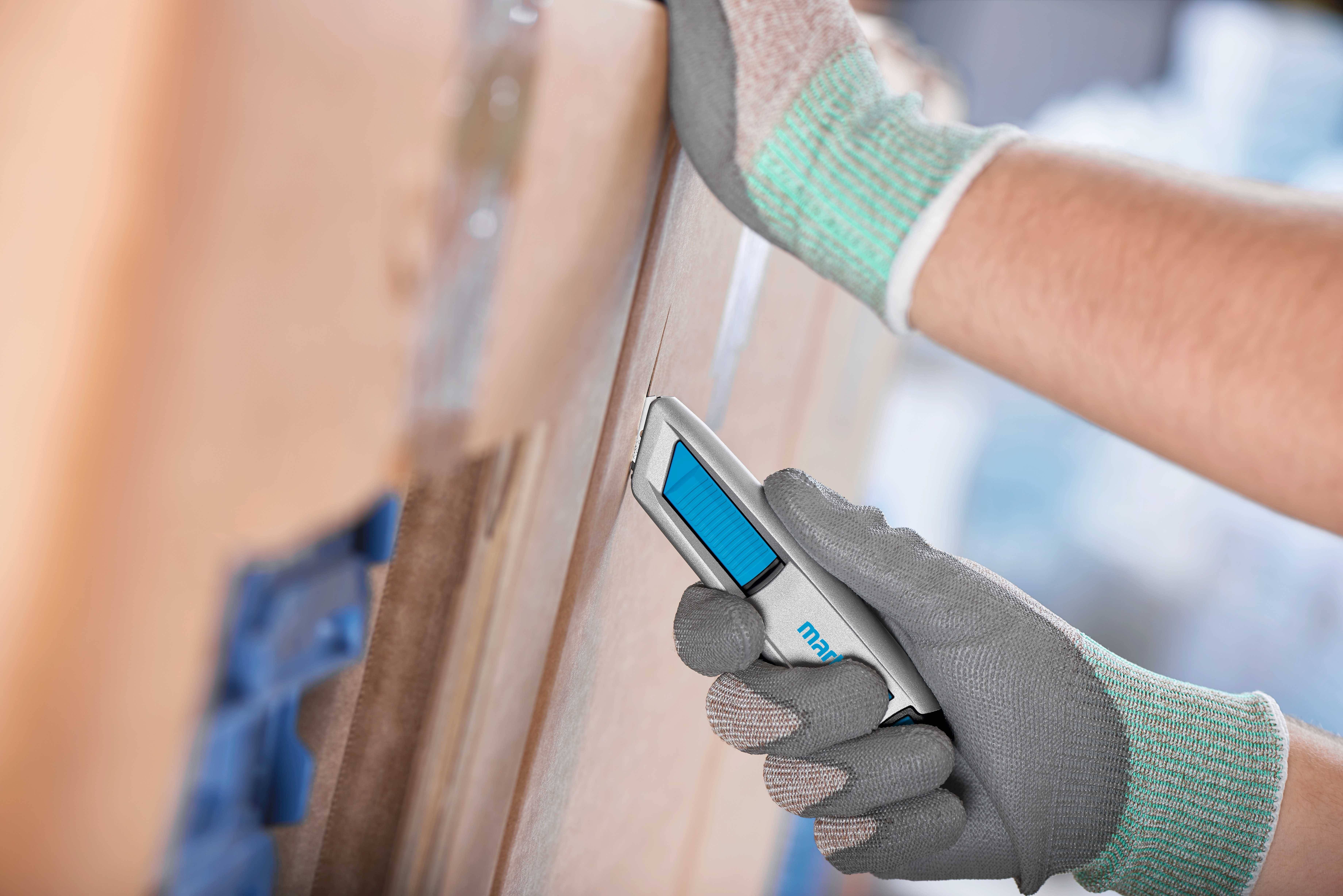 A person wearing work gloves is using a safety knife to open a cardboard box. The environment suggests a warehouse setting.