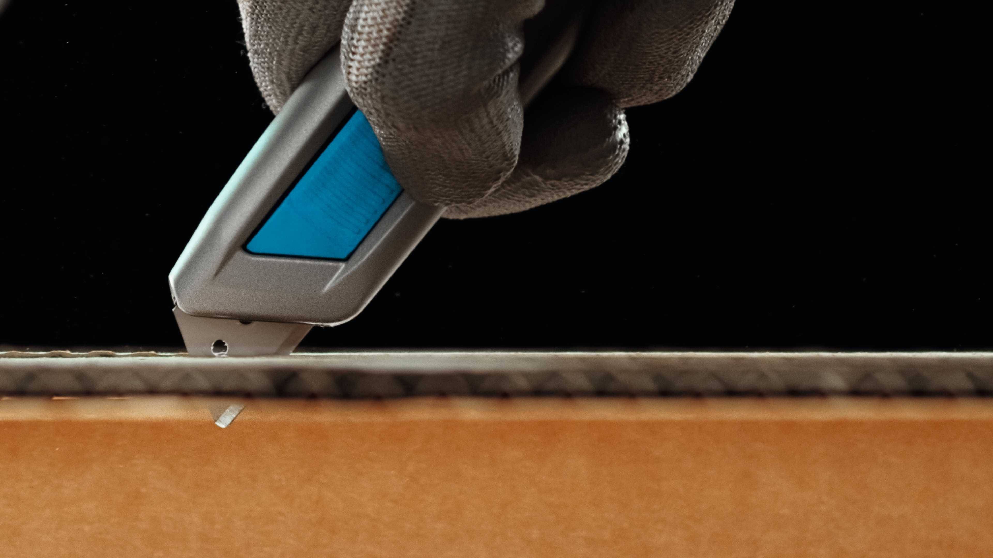 A hand wearing a glove cuts cardboard with a Stanley knife.