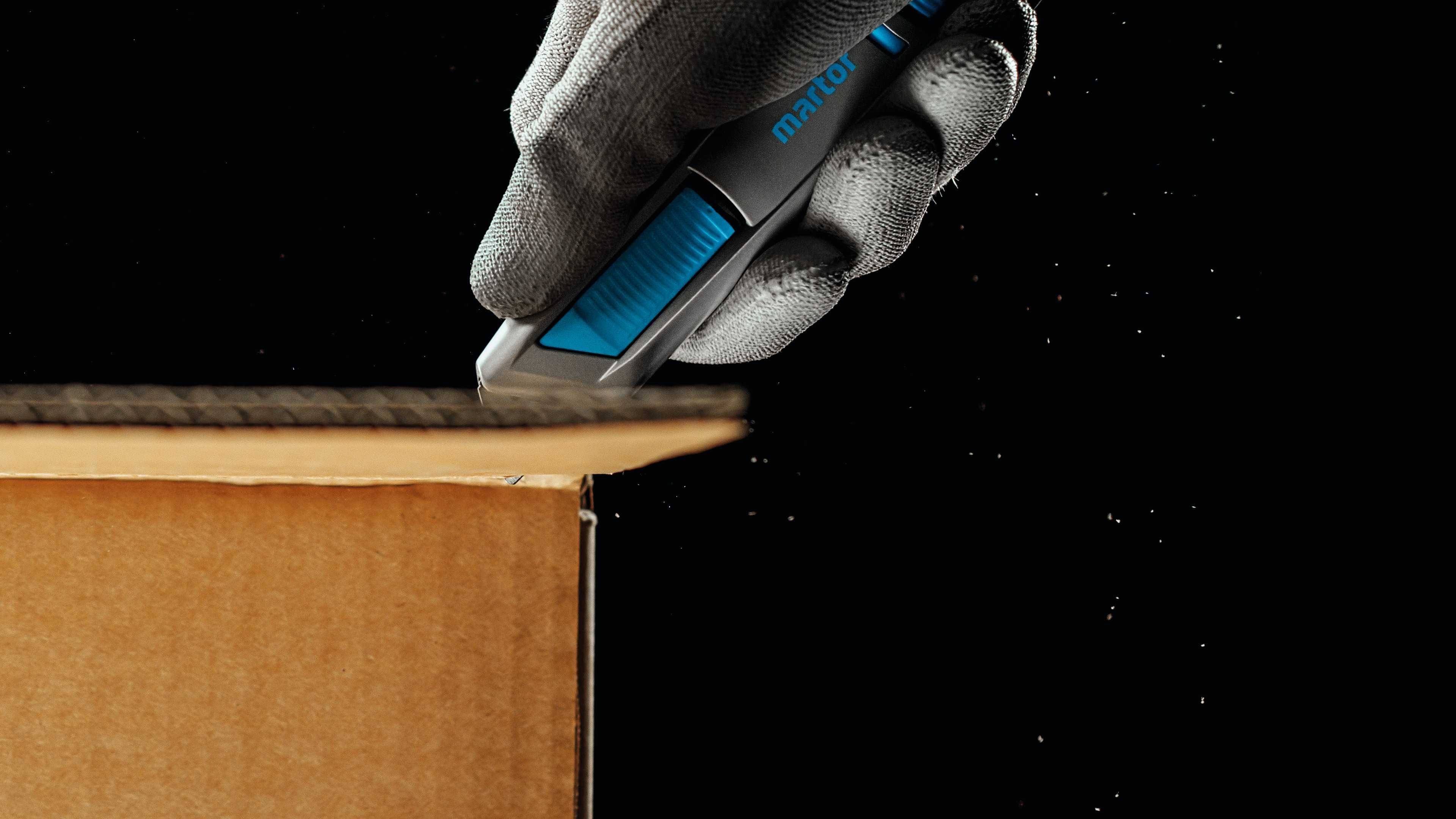 A gloved hand carefully cuts the top of a cardboard box with a blue utility knife against a black background.