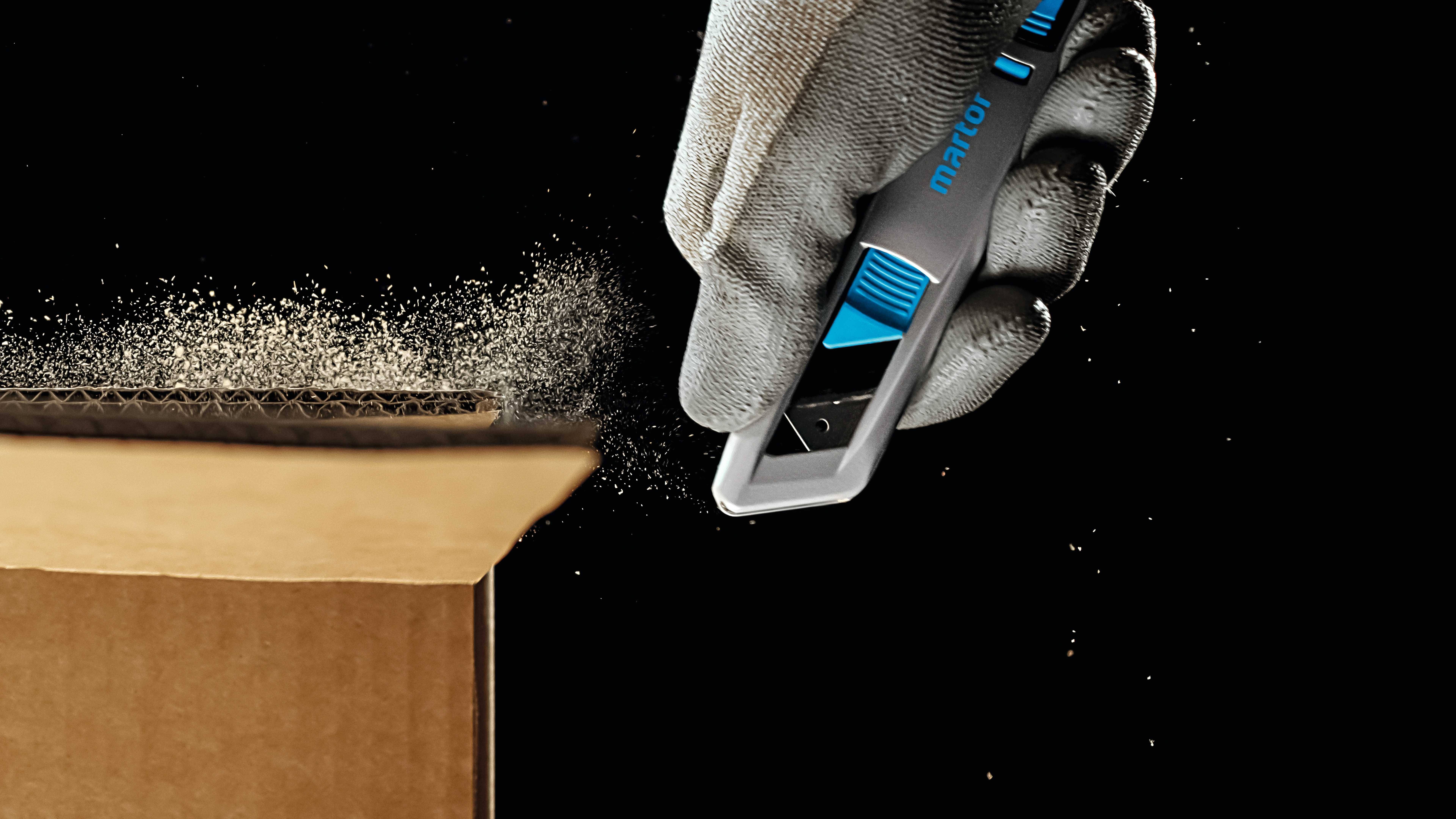A gloved hand precisely cuts through a cardboard box with a grey knife. Dust particles hover in the air.