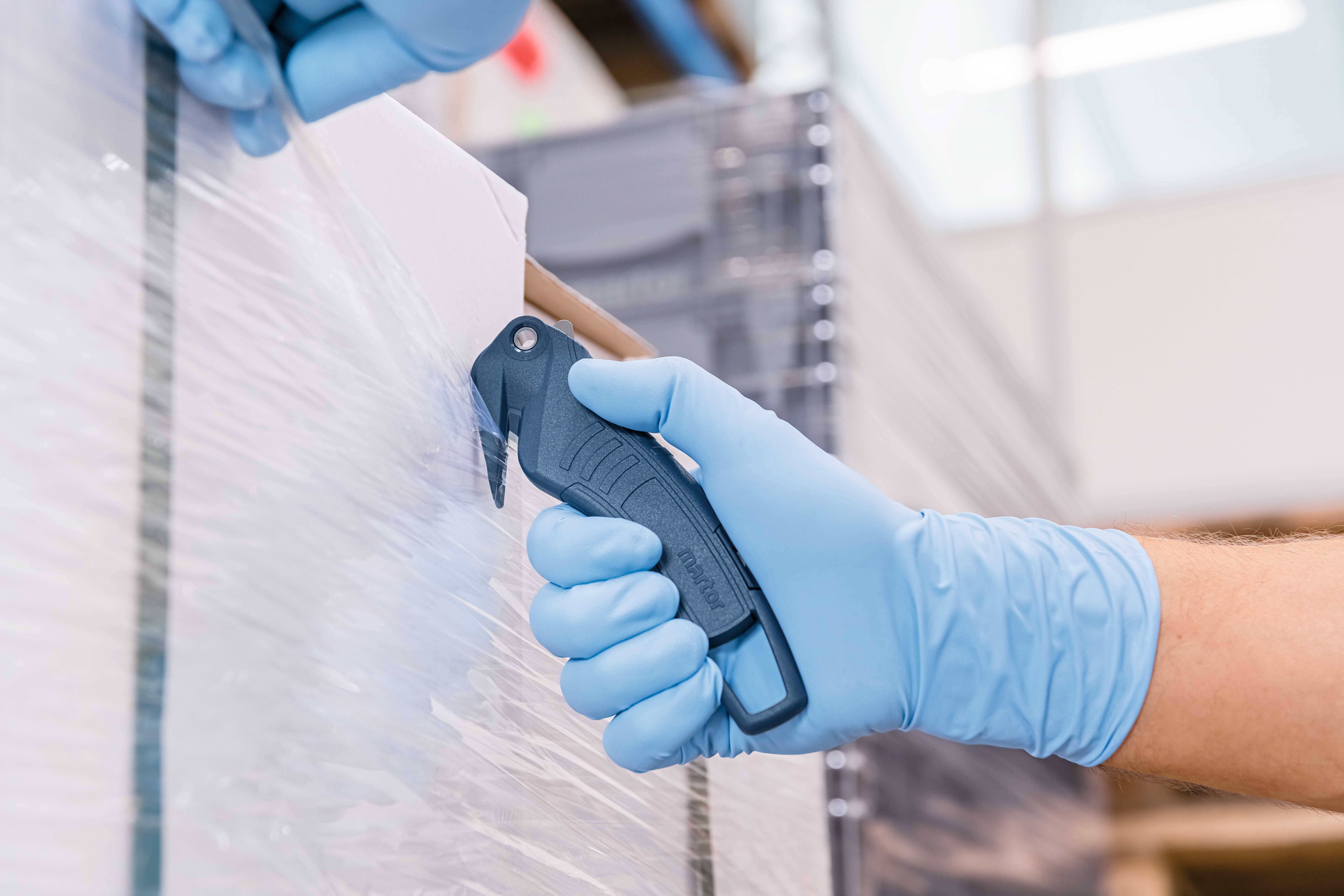 'Two hands wearing blue gloves are cutting plastic film with a safety knife from a pallet to expose the goods.'
