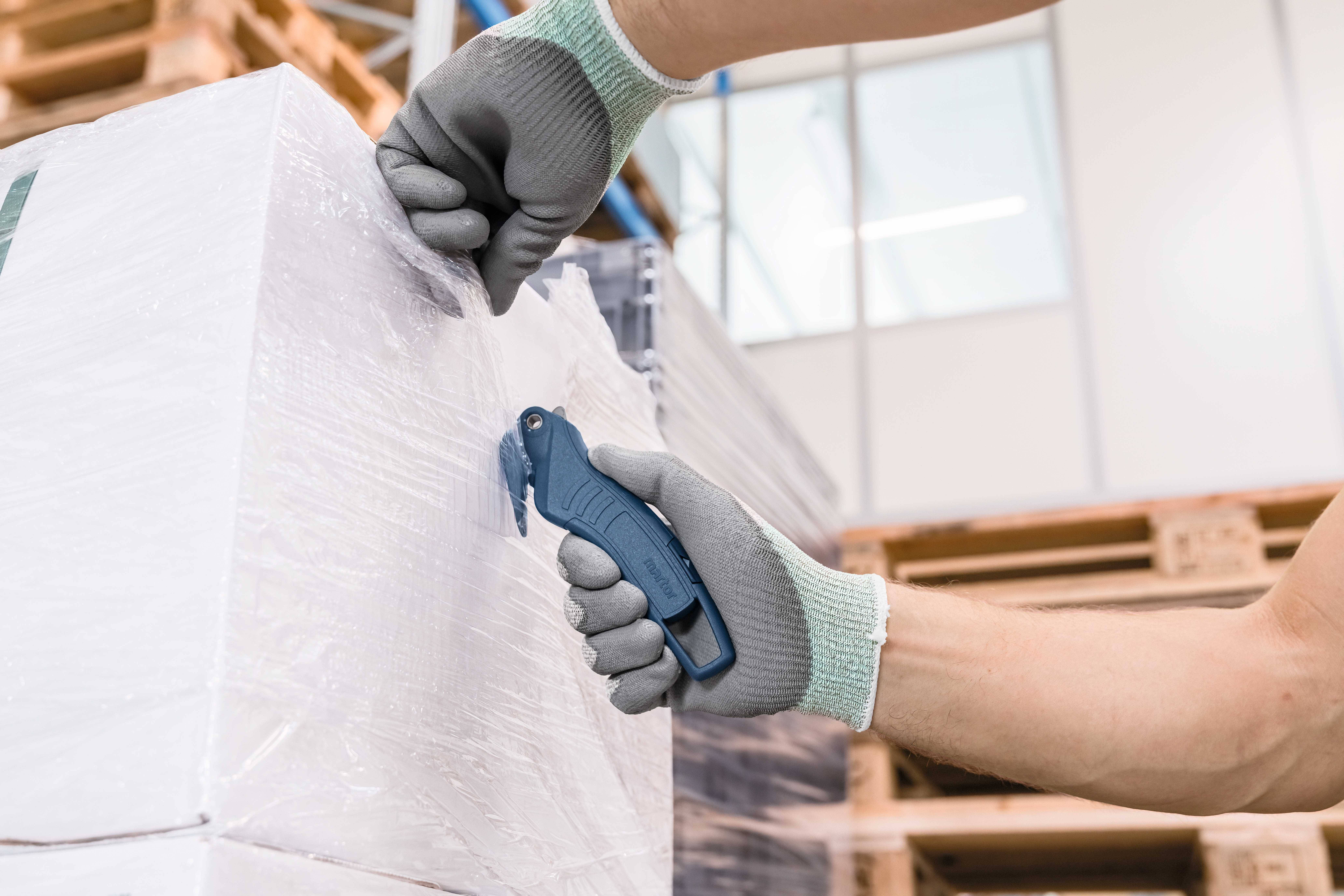 A person wearing grey gloves is cutting plastic film from a pallet in the warehouse using a blue safety cutter.