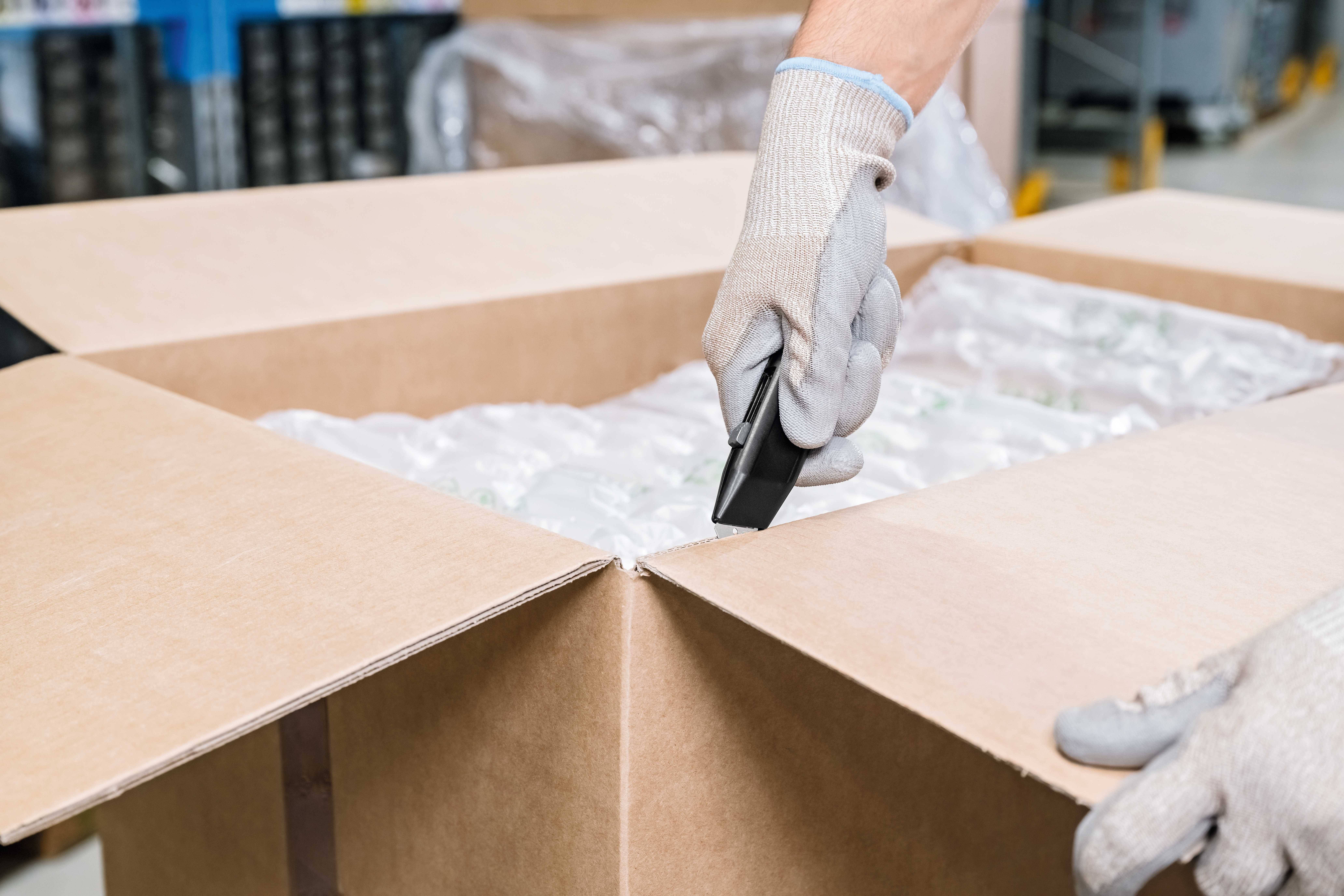 A person opens a large cardboard box filled with packaging material using a Stanley knife.