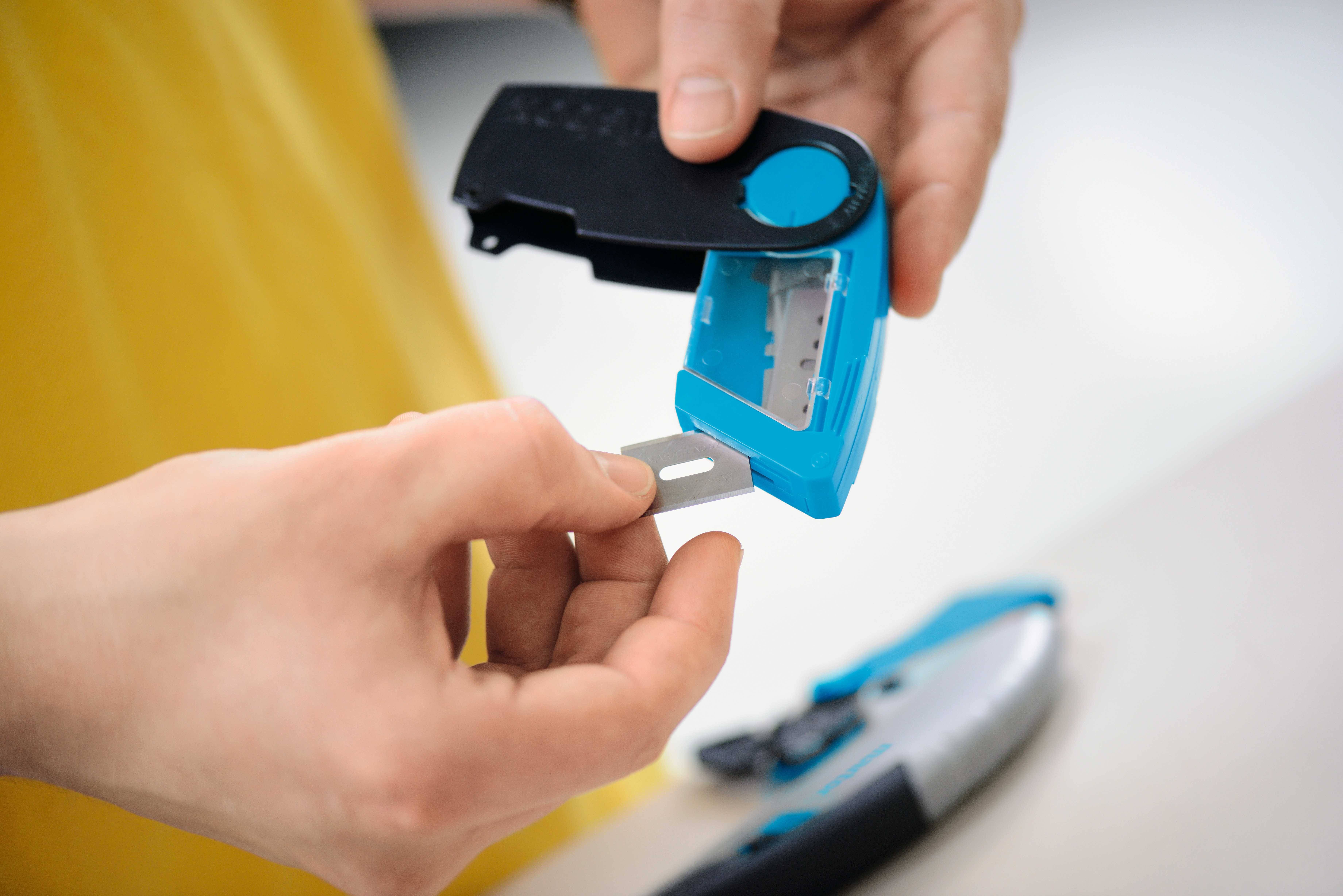A person opens a casing to insert a replacement blade for a blue utility knife. Their hands are holding the blade and the casing.