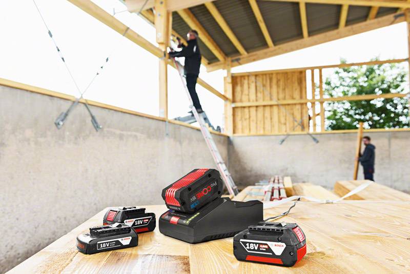 Four Bosch batteries and a charger on a wooden table at a construction site; in the background, a person is working on a ladder.
