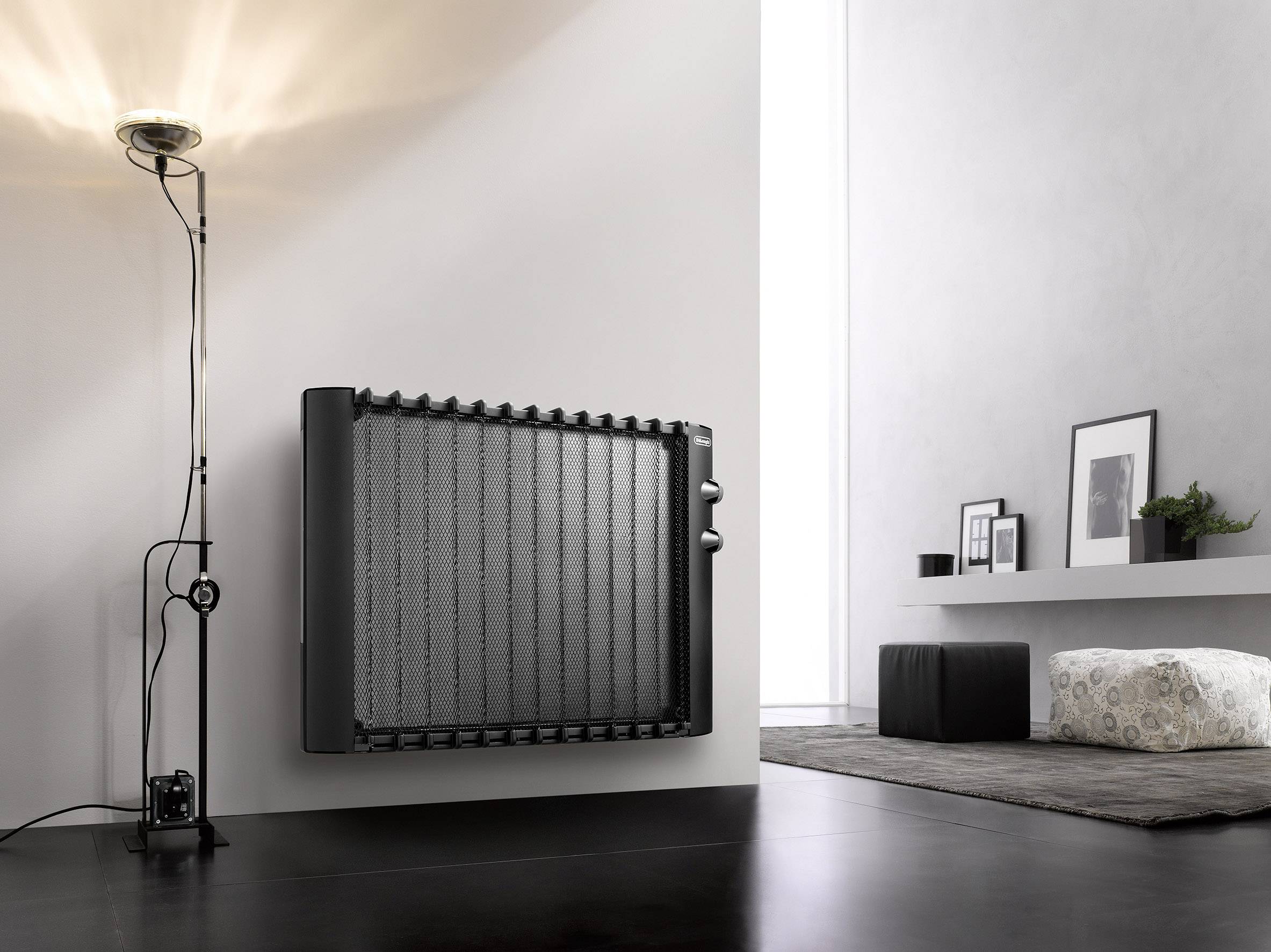 A contemporary room with a black heater mounted on the wall. A standard lamp illuminates the area; beside it are some scatter cushions and artwork.