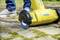 Someone is cleaning a paved surface with a yellow electric brush. The brush is removing moss from between the stones.