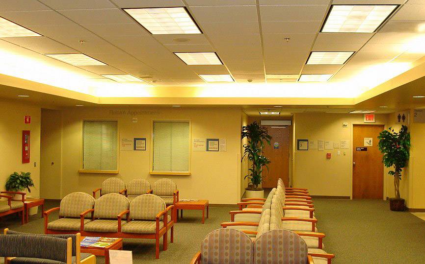A waiting room with beige walls, upholstered chairs, wooden tables and plants. Bright ceiling lights illuminate the space.