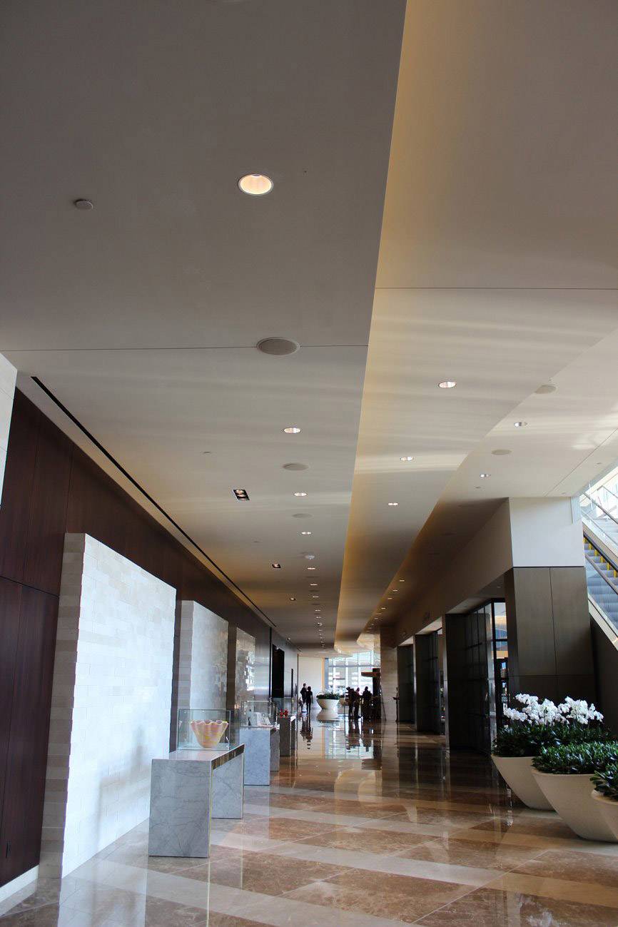 An elegant hotel corridor with a marble floor. On the left are decorative elements, while on the right are large windows and plant arrangements.
