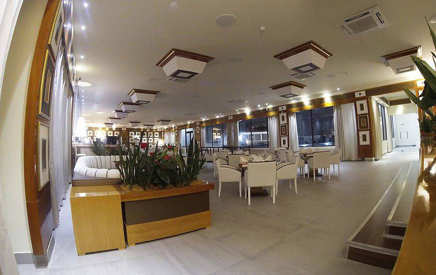 An elegant restaurant with contemporary decor. White tables and beige chairs are positioned beside large windows. Plants divide the seating areas.