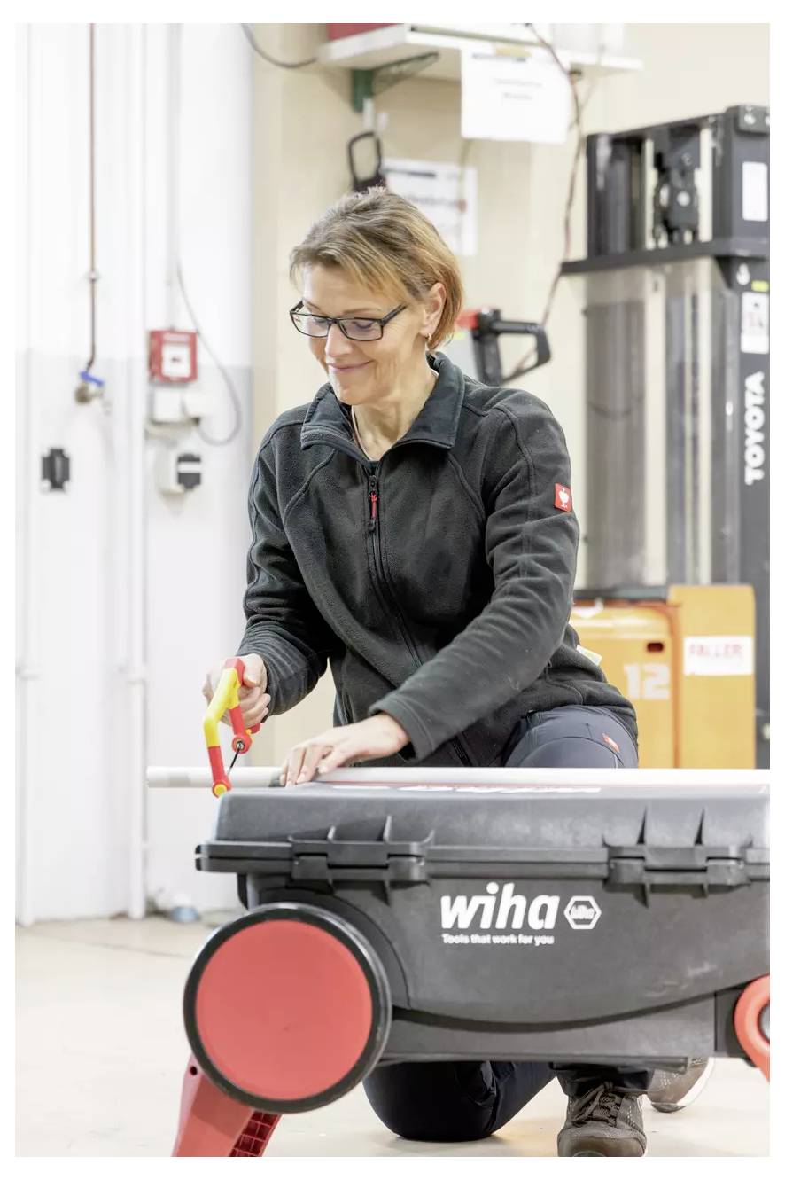 A person wearing a jacket is using a tool on a workbench with wheels. Industrial equipment and a fire alarm are visible in the background.