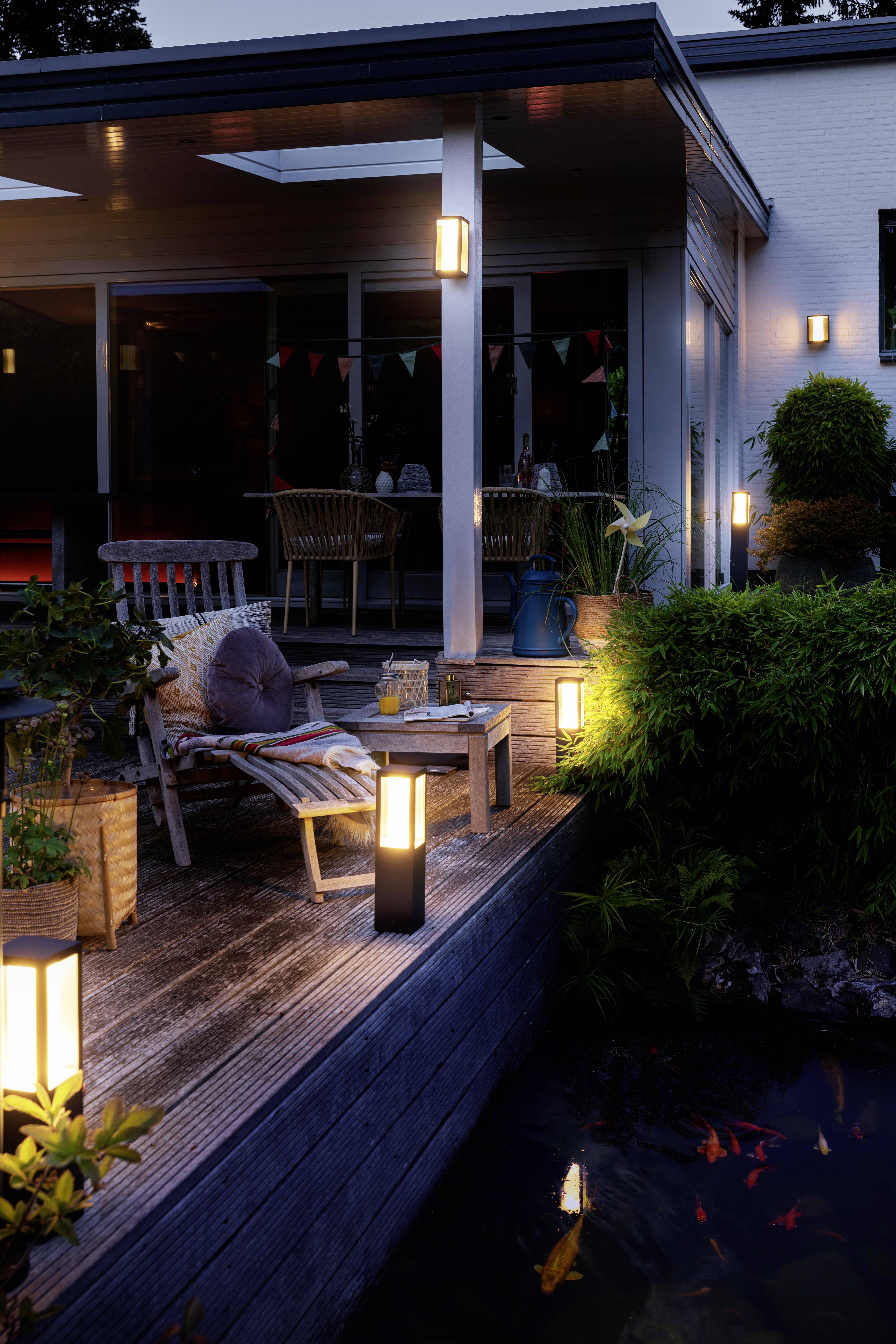 An evening terrace with lighting. Lounge furniture and a table are positioned on the wooden deck. In the foreground, a pond with fish is visible.