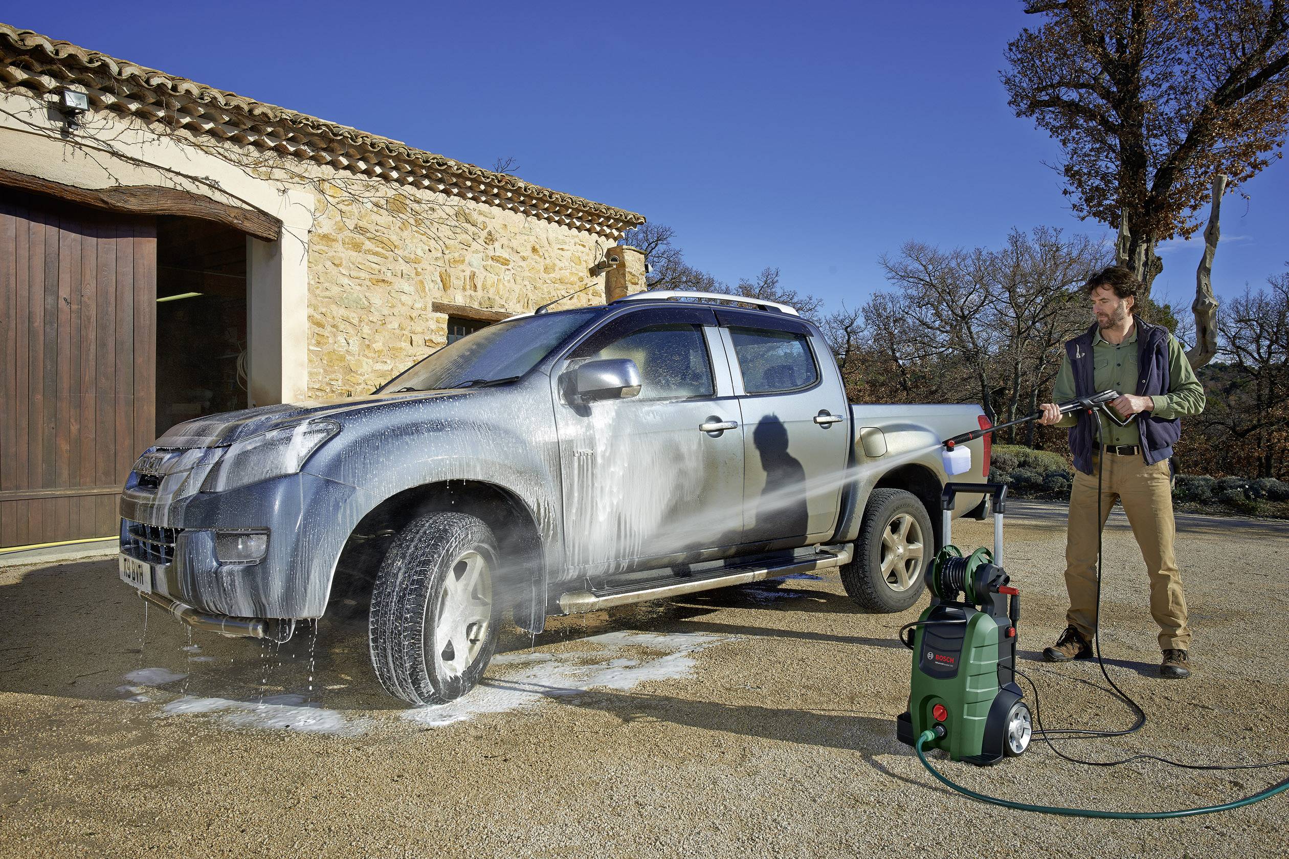 A man is cleaning a pickup truck with a high-pressure washer in the yard of a stone building. The vehicle is covered in soapy water.