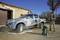 A man is cleaning a pickup truck with a high-pressure washer in the yard of a stone building. The vehicle is covered in soapy water.