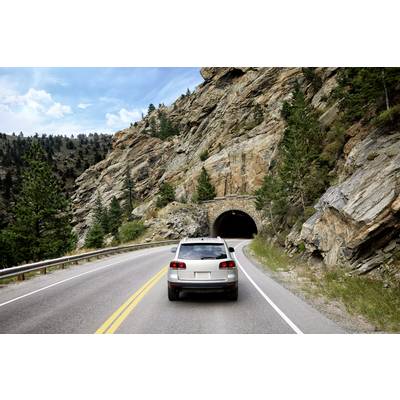 A car is driving on a road through a rocky landscape, with a tunnel in the background, surrounded by trees and a blue sky.