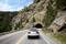 A car is driving on a road through a rocky landscape, with a tunnel in the background, surrounded by trees and a blue sky.
